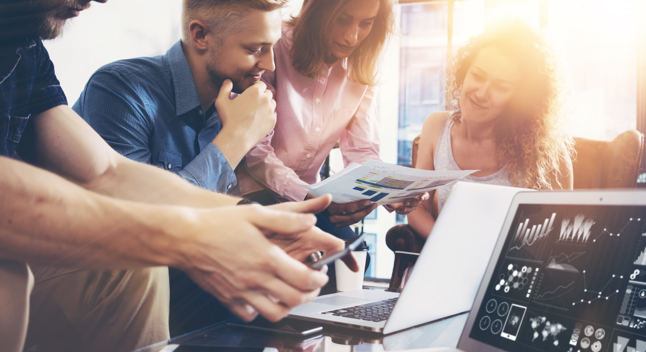 A group of people engaging in a discussion around documents and laptops in a sunlit office, conveying teamwork and collaboration