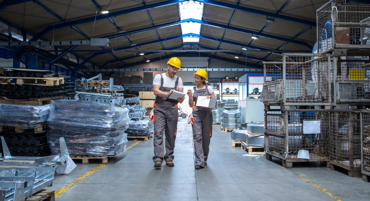 Two workers in a warehouse wearing yellow helmets and gray uniforms are walking and discussing while holding a clipboard and tablet